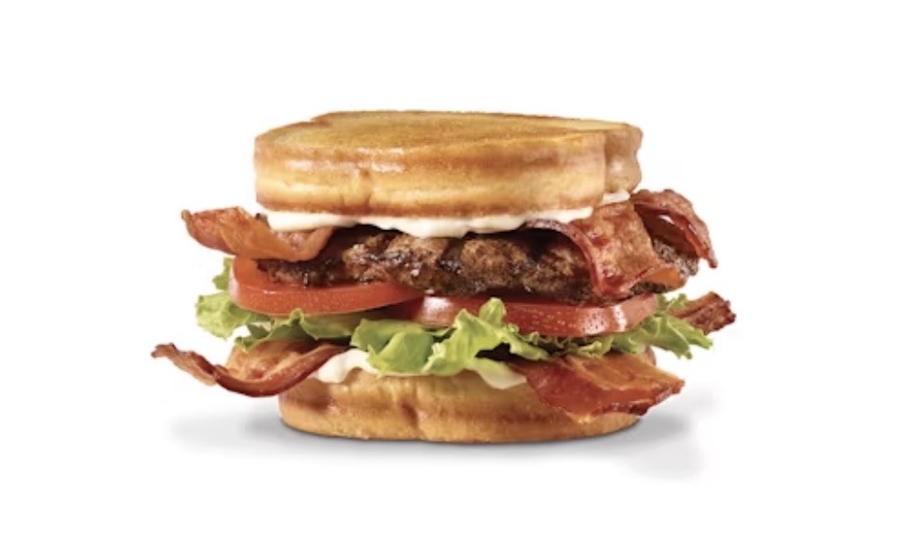 A delicious new Hardee's sandwich featuring crispy bacon, lettuce, tomato, a burger patty, and mayonnaise, all served between two golden brown waffle buns on a white background.
