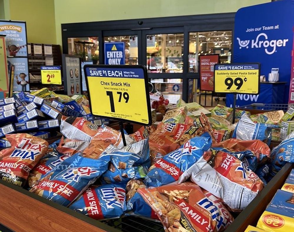 Bags of Chex Snack Mix are displayed for sale in a grocery store with a yellow $1.79 price sign. A sign for Suddenly Salad pasta salad priced at 99 cents is visible in the background near the entrance.