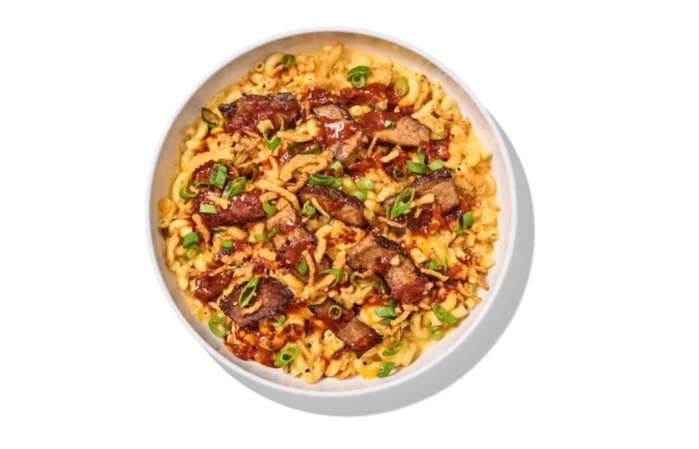 A white bowl filled with macaroni and cheese, topped with pieces of grilled meat, chopped green onions, and a drizzle of barbecue sauce—a delicious creation from the new fast-food scene.