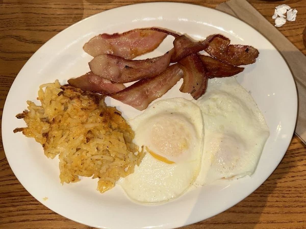 A plate with two strips of bacon, two fried eggs, and a serving of hash browns sits on a wooden table at Cracker Barrel. A napkin and a crumpled piece of paper are nearby.