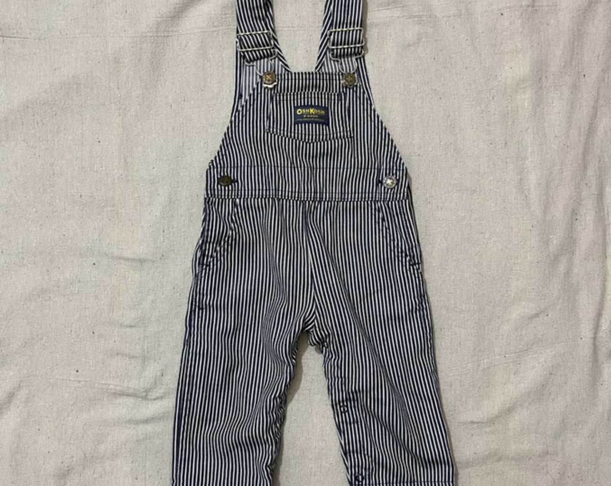 A pair of children's blue-and-white striped overalls with adjustable shoulder straps, metal clasps, and a front pocket, styled in a classic 1980s look, laid flat on a plain light fabric background.
