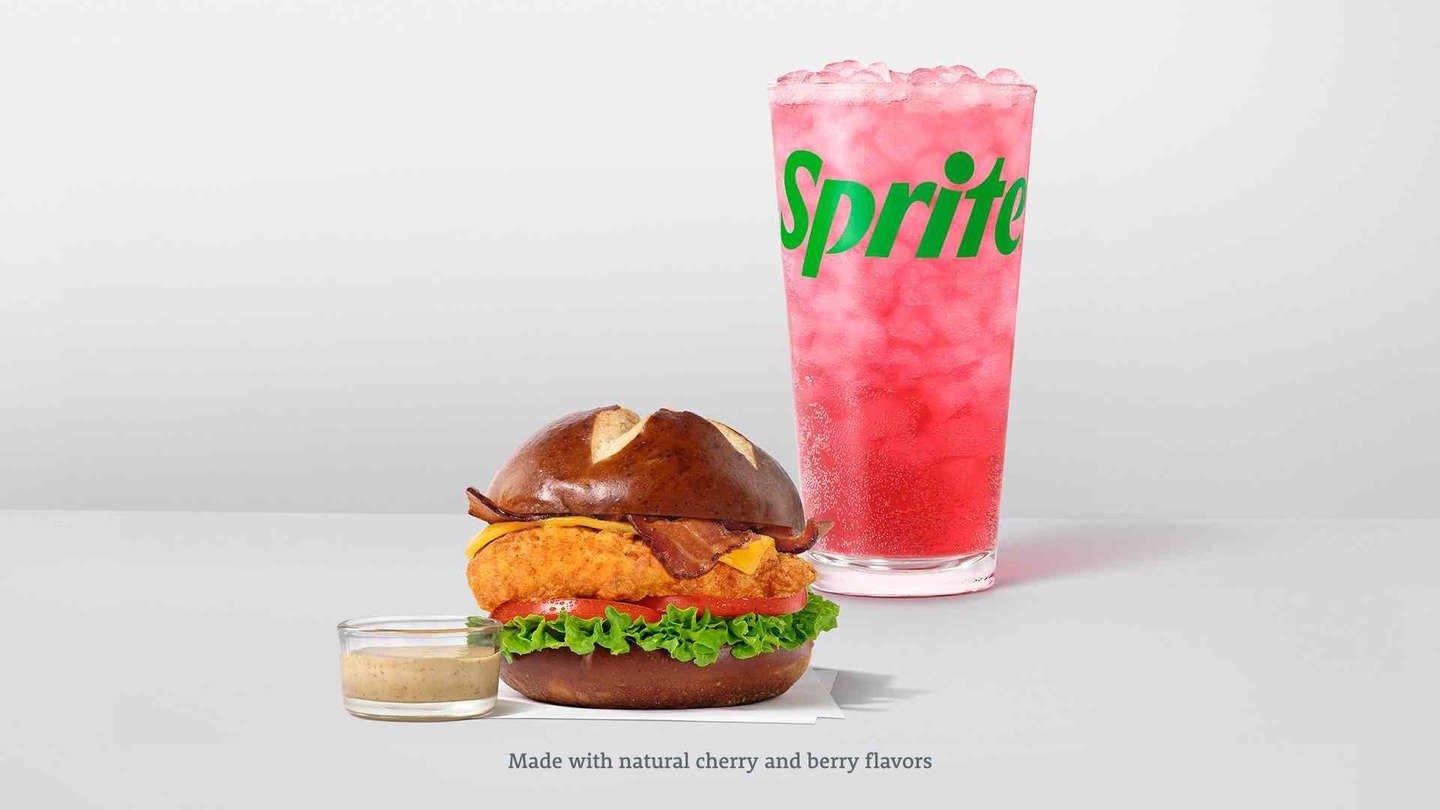 A new fast-food sandwich with lettuce, bacon, cheese, and a fried chicken patty in a pretzel bun sits beside a cup of pink Sprite with ice. A small cup of dipping sauce is placed next to the sandwich.