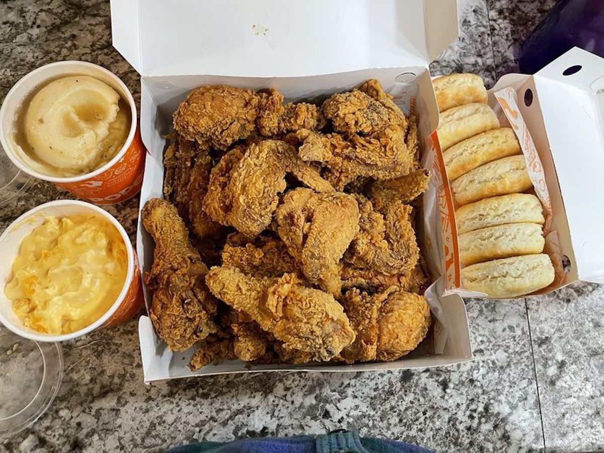 A box of fried chicken pieces, a side of mashed potatoes, a side of macaroni and cheese, and a box of biscuits are arranged on a granite countertop.
