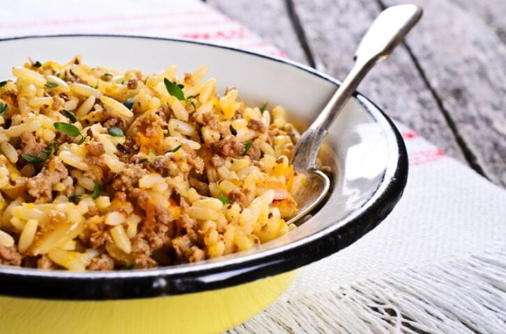 A close-up of a bowl filled with a rice dish mixed with ground meat, herbs, and spices. A spoon rests inside the bowl, which is placed on a fringed white cloth on a rustic wooden table.