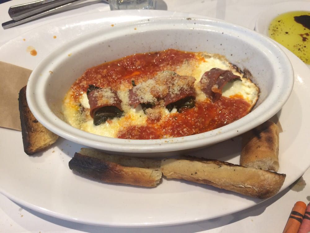 A white oval dish with baked eggplant rollatini topped with tomato sauce and grated cheese, surrounded by toasted bread sticks on a white plate. A small dish of olive oil with herbs is partially visible on the side.