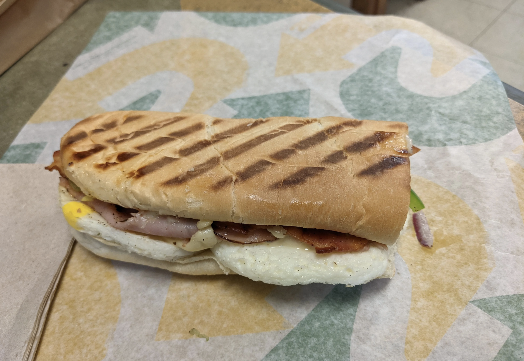 A toasted sub sandwich with grill marks, filled with egg, bacon, ham, cheese, and vegetables, sits on branded sandwich paper.