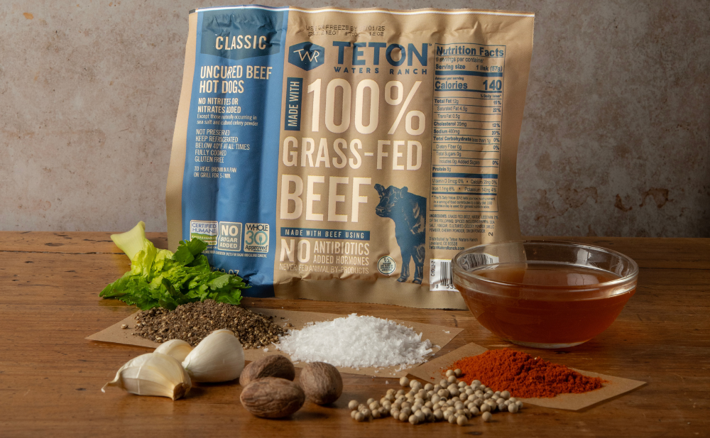 A package of Teton Waters Ranch uncured beef hot dogs stands behind a wooden table with seasonings, garlic, nutmeg, paprika, whole peppercorns, salt, fresh herbs, and a small bowl of brown broth arranged in front.
