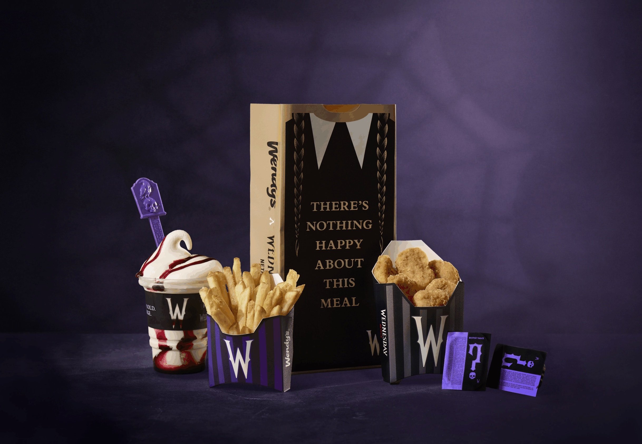 A new fast-food Wendy’s meal featuring fries, chicken nuggets, a sundae, and special gothic-style packaging with "There's nothing happy about this meal" on the box, all set against a dark purple background.