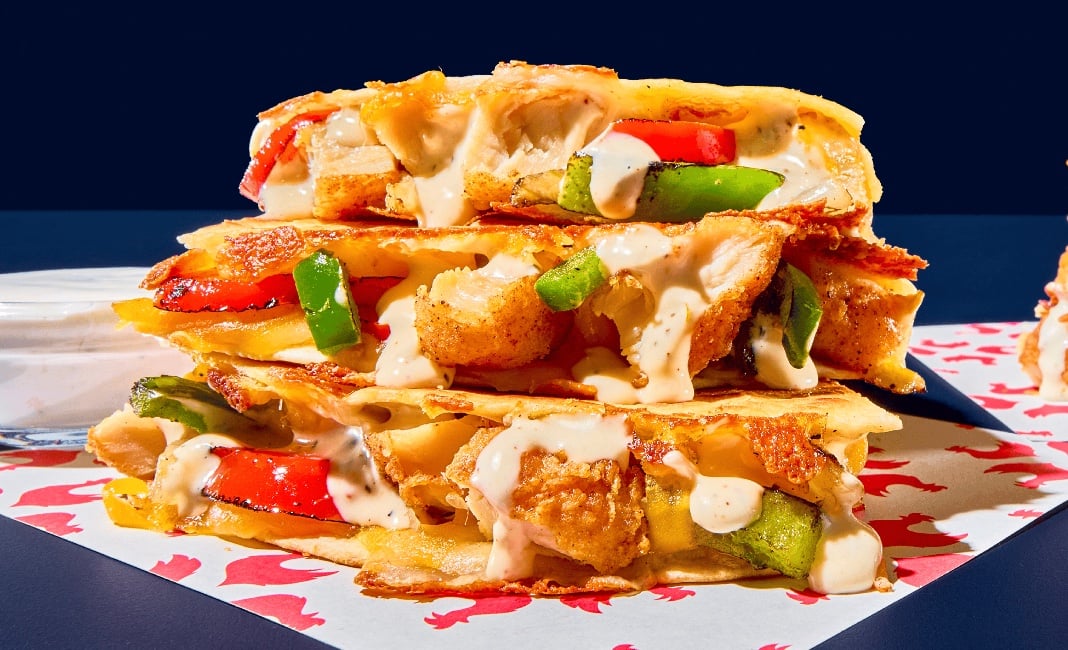 A stack of cheesy quesadilla wedges filled with grilled chicken, red and green bell peppers, and drizzled with creamy sauce brings the best of new fast-food trends, displayed on patterned paper against a dark background.