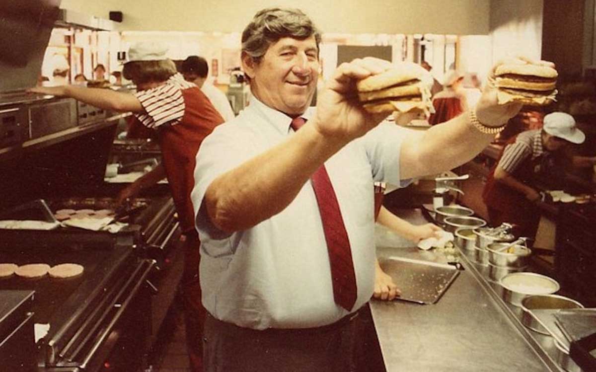 A man in a shirt and tie smiles while holding up two burgers in a busy fast food kitchen, with other workers preparing food in the background.