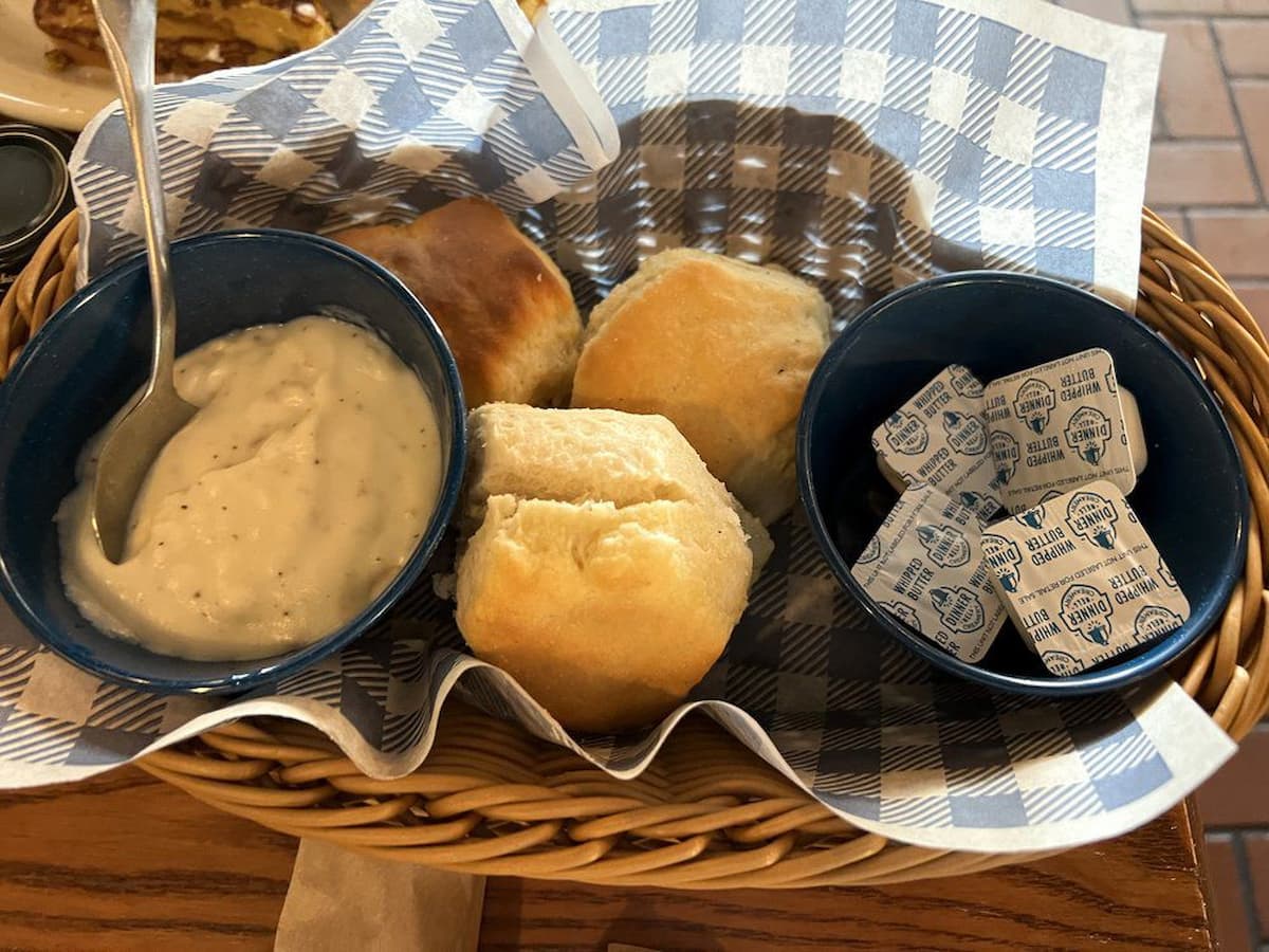A wicker basket with blue and white checkered paper holds three biscuits, a bowl of white gravy with a spoon, and a bowl with several packets of salted butter—just like you'd find at Cracker Barrel.