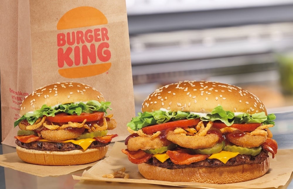 Two Burger King sandwiches with sesame seed buns, lettuce, tomato, pickles, crispy bacon, onion rings, cheese, and beef patties are shown—a delicious new fast-food option—with a Burger King paper bag in the background.