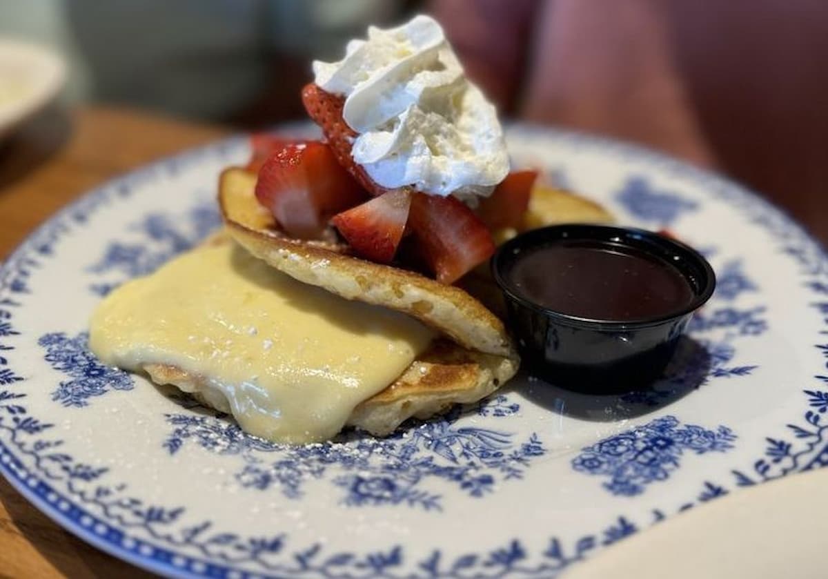 A blue and white floral plate with pancakes topped with melted cheese, fresh strawberries, whipped cream, and a small cup of syrup on the side—just like a cracker barrel breakfast classic.