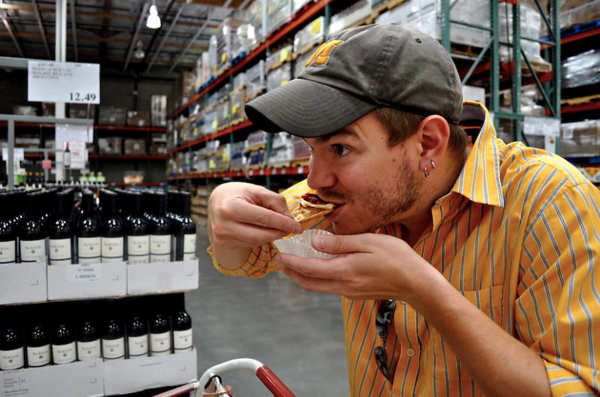 A man wearing a striped shirt and cap samples food in a large warehouse store, standing near shelves stocked with wine bottles and other goods—a scene familiar to those seeking Costco shopping tips.