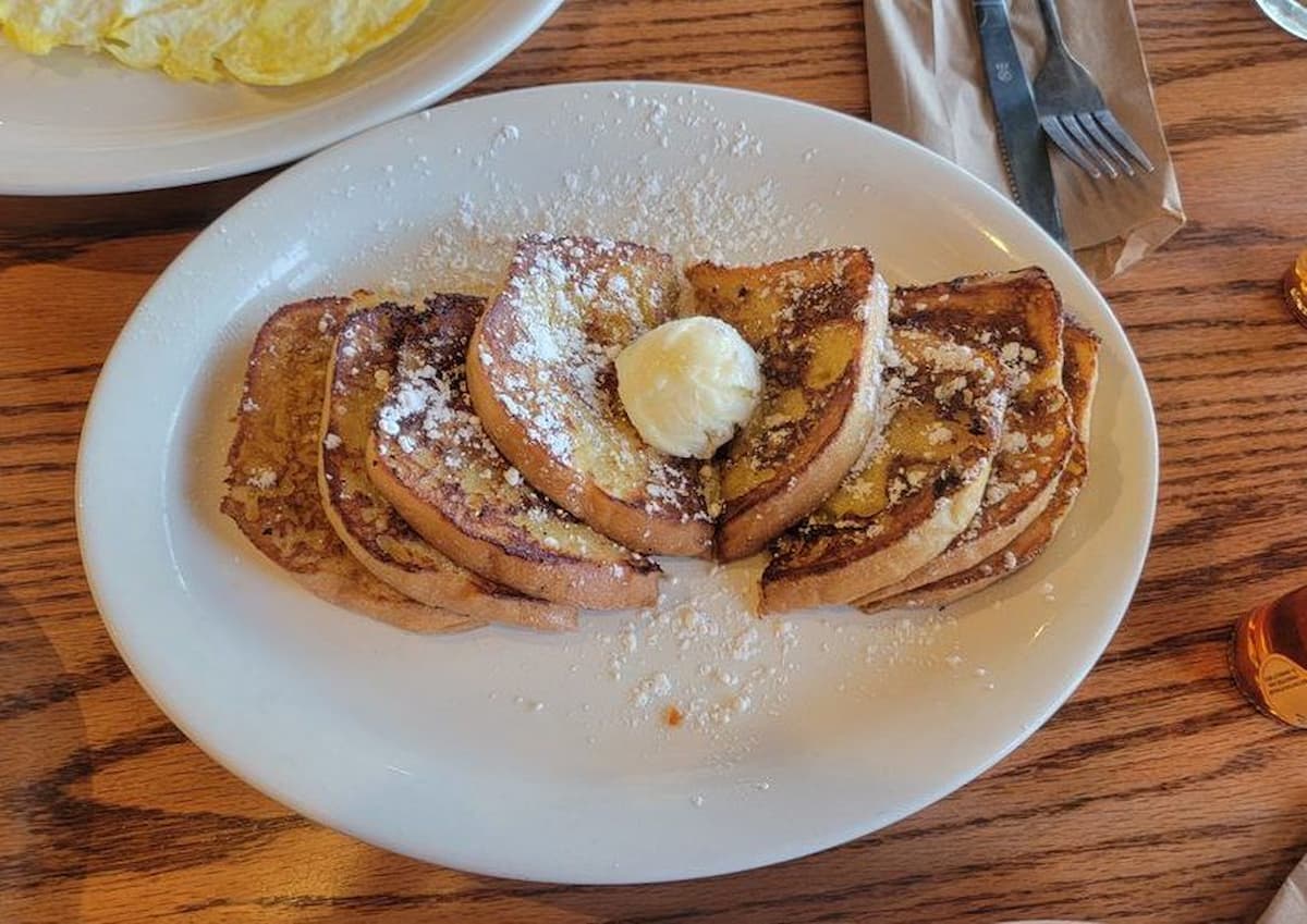 Six slices of French toast topped with powdered sugar and a dollop of butter are arranged on a white plate, capturing the classic comfort of a Cracker Barrel breakfast, with another plate and utensils visible in the background.