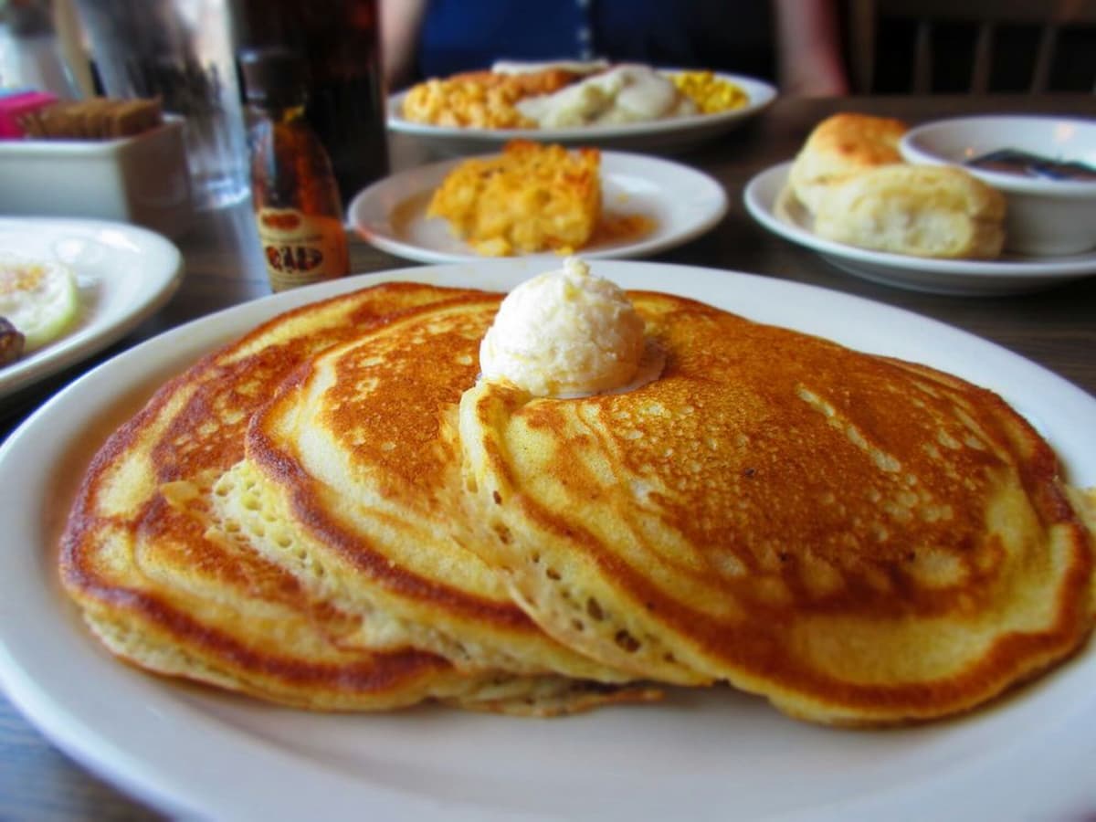 Three golden pancakes topped with a dollop of butter are served on a white plate, just like at Cracker Barrel, with other breakfast dishes such as biscuits, eggs, and a drink visible in the background.