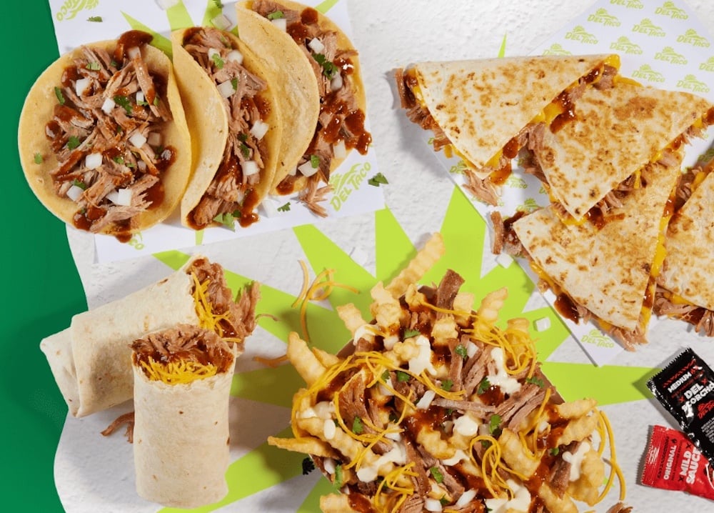 An overhead view of new fast-food Del Taco Mexican favorites: tacos, quesadillas, burritos, and loaded fries with shredded meat, cheese, sauce, and fresh herbs, served alongside hot sauce packets on a white and green background.