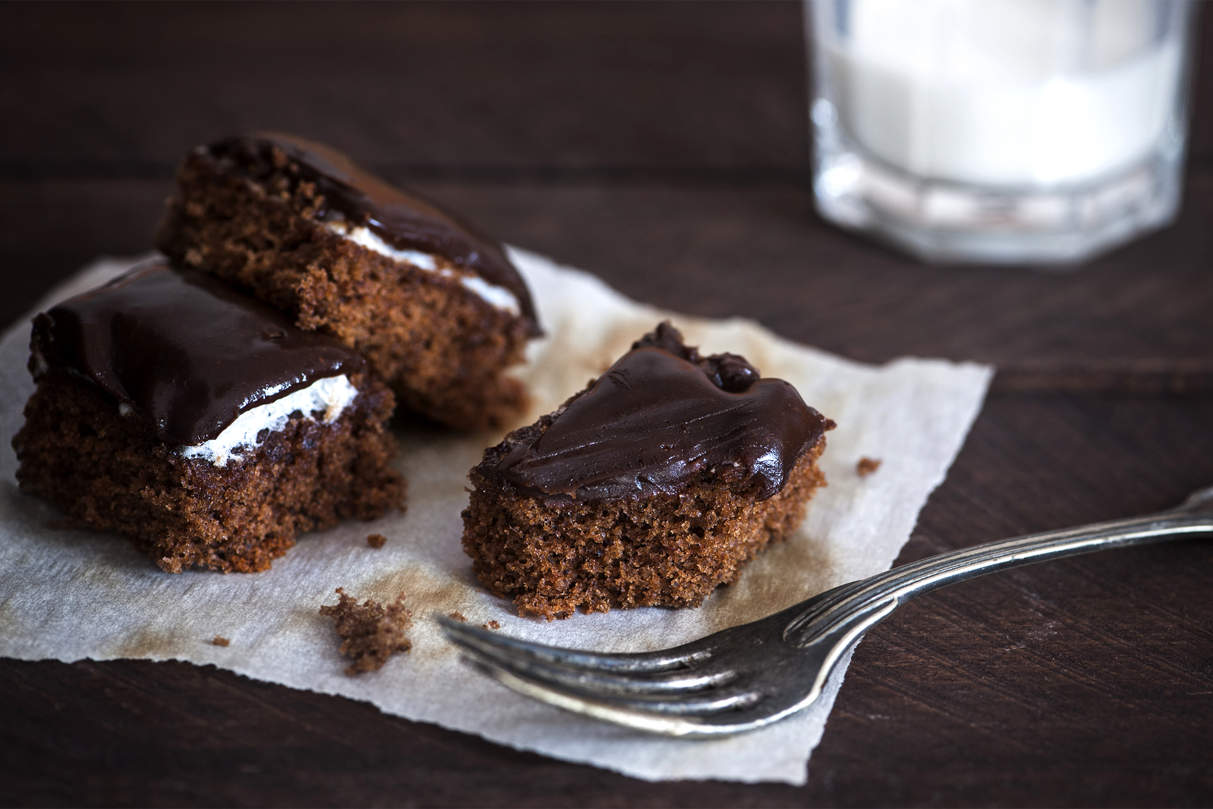 Three pieces of chocolate cake with glossy chocolate frosting and cream filling sit on parchment paper beside a fork. A glass of milk is blurred in the background on a dark wooden surface.