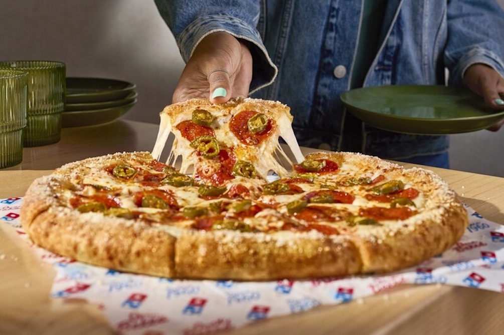 A person in a denim jacket lifts a cheesy slice of pepperoni and jalapeño pizza from a whole pizza on Domino's branded paper, with stacked green plates and glasses in the background, capturing the vibe of new fast-food favorites.