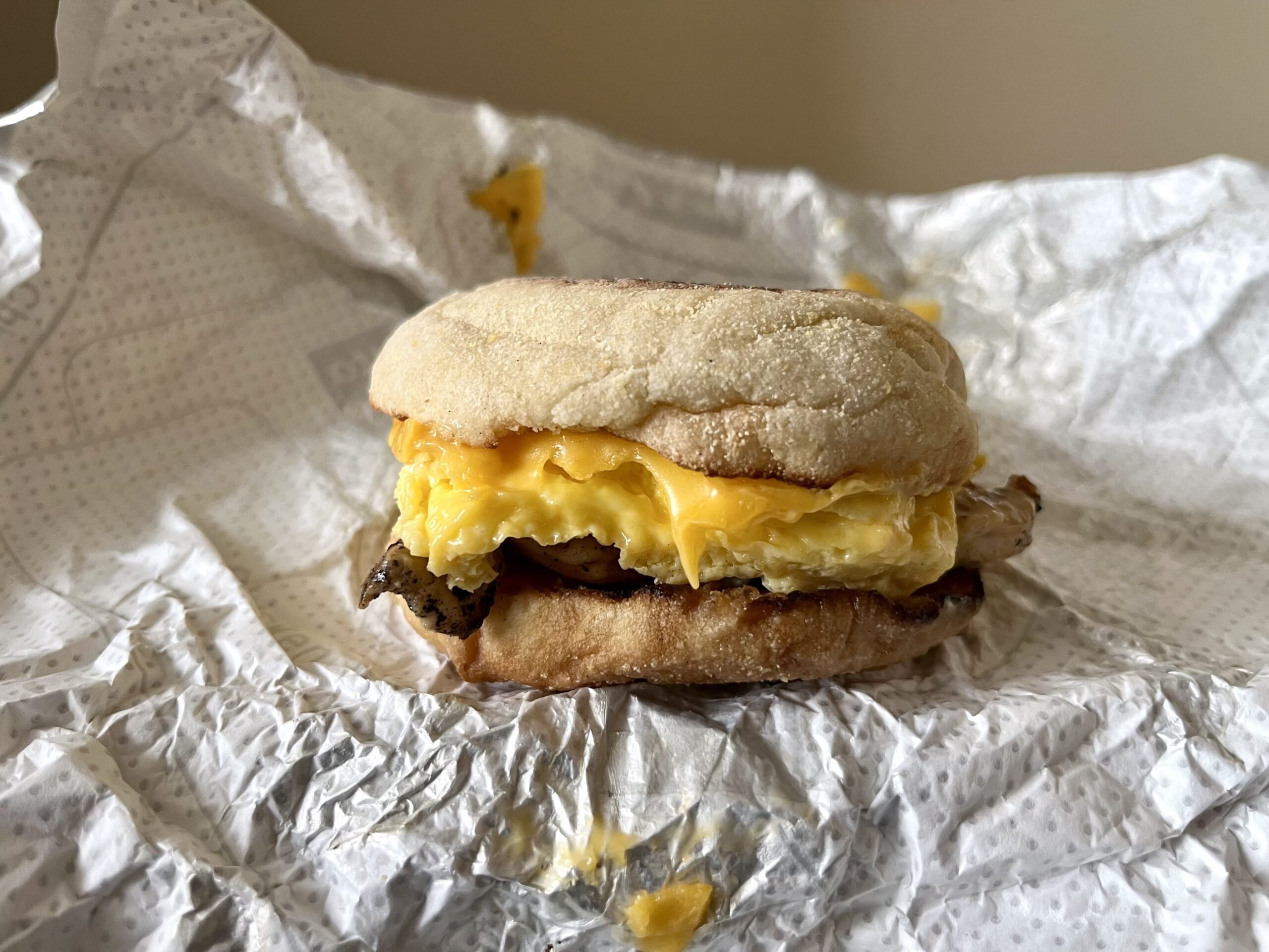 A breakfast sandwich with scrambled eggs, melted cheese, and what appears to be sausage, served on an English muffin, sits on a crumpled white wrapper.