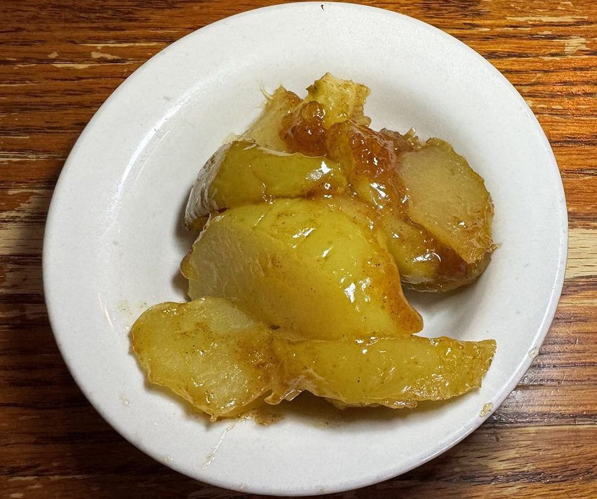 A small white plate holds several slices of glazed baked apples on a wooden table, reminiscent of Cracker Barrel’s classic comfort food. The apple slices are shiny and coated in a syrupy sauce.