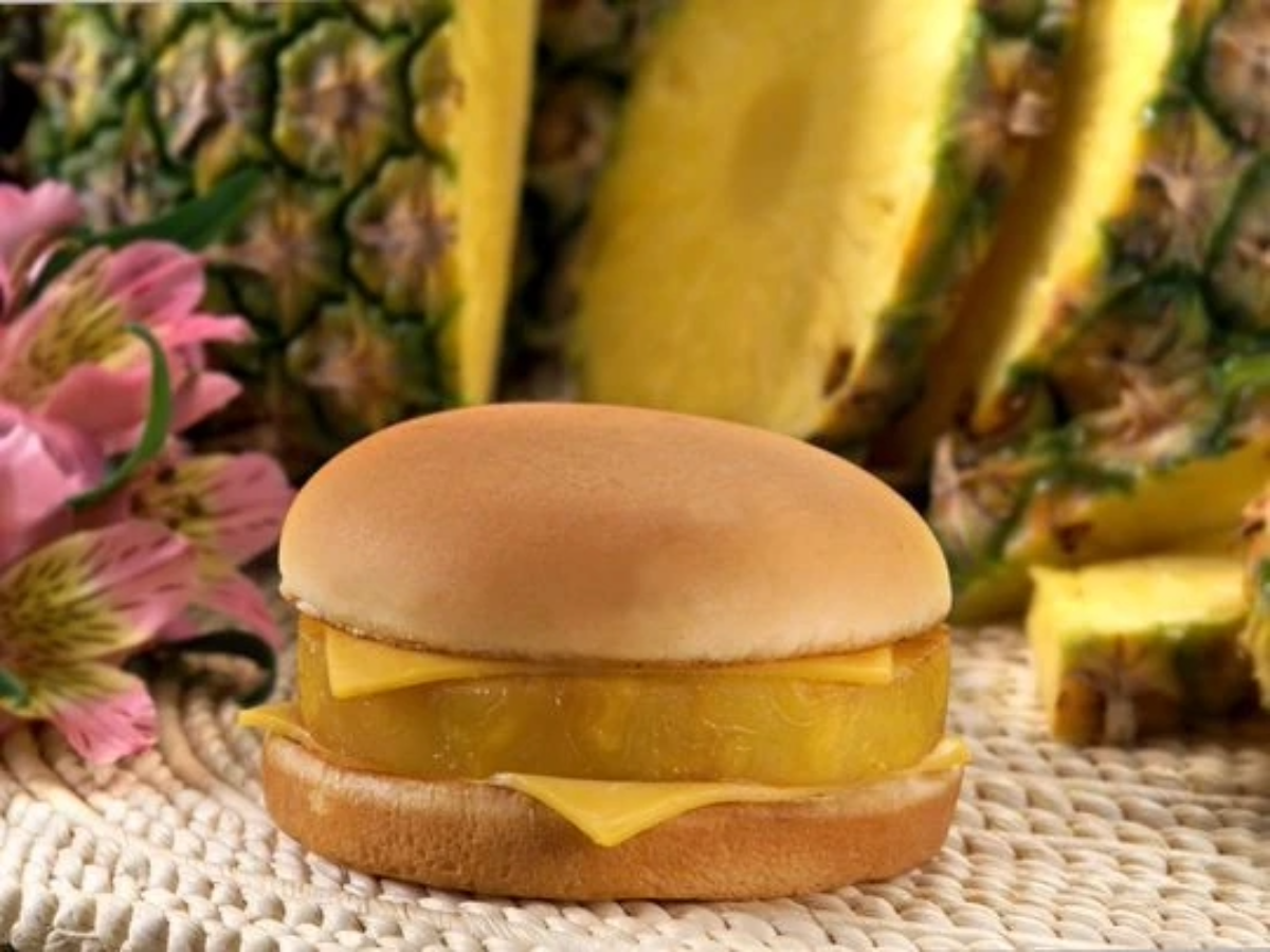 A sandwich with a round bun, a slice of cheddar cheese, and a thick slice of pineapple, set on a woven mat with pineapple slices and pink flowers in the background.