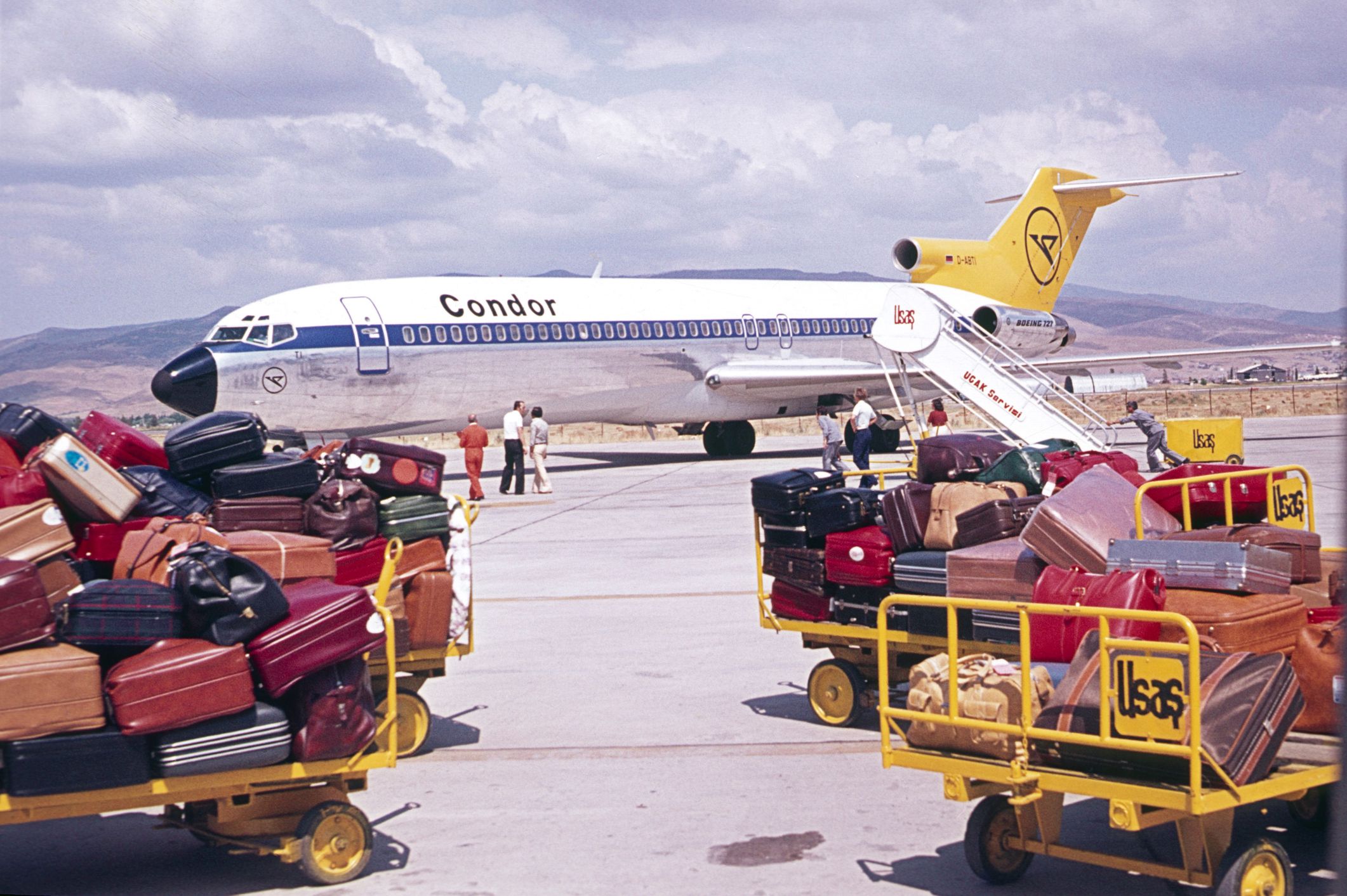 Andalusia, Spain, 1976. Mass tourism. Tourist luggage on an airfield. In the background a charter plane.