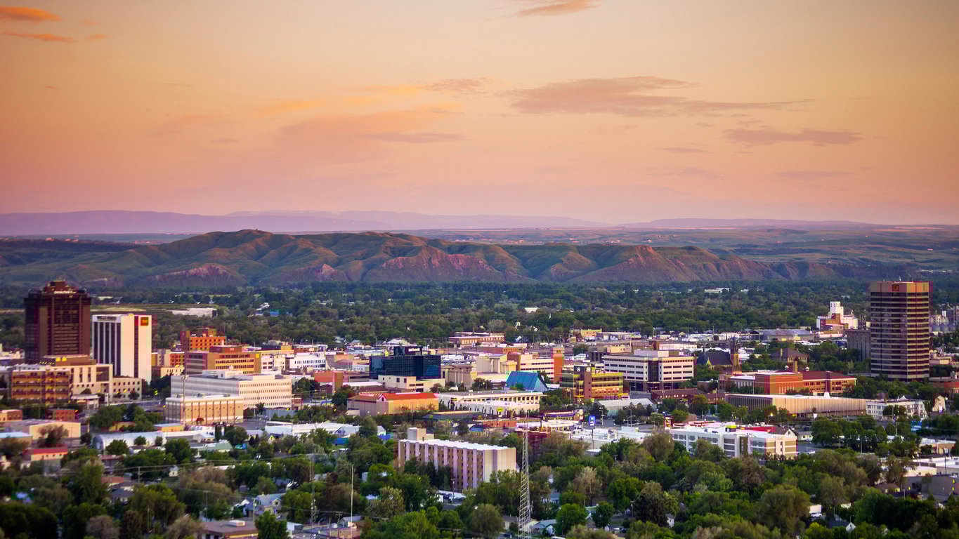 Downtown Billings, Montana at sunset, with city buildings in the foreground and green hills and distant mountains under a pastel sky in the background.