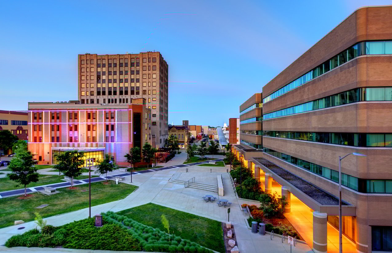 A modern cityscape shows a campus setting with colorful lights illuminating a building on the left, adjacent to a tall office tower, and a multi-story building on the right, all under a clear evening sky.