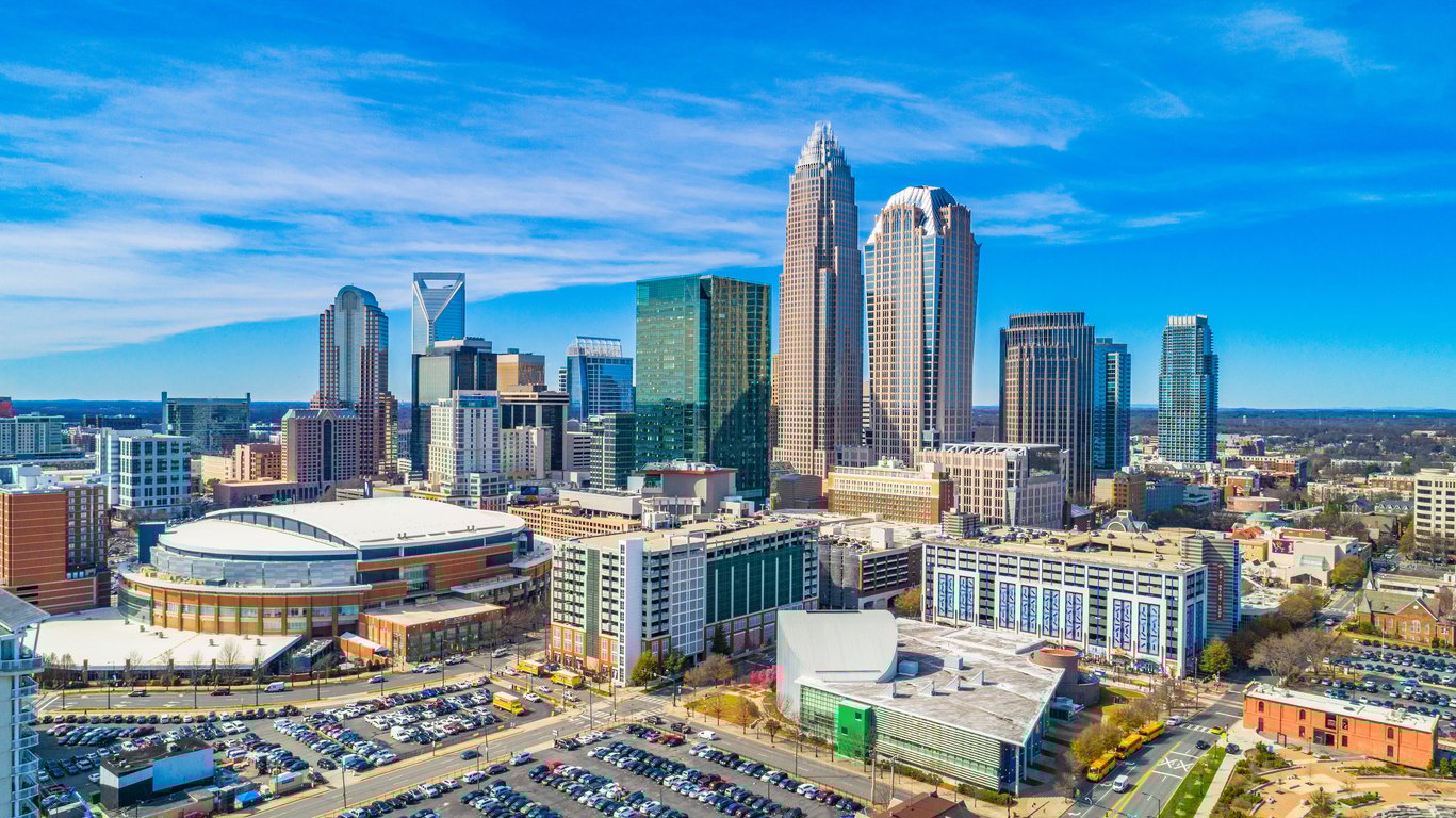 Aerial view of downtown Charlotte, North Carolina, showing modern skyscrapers under a blue sky, with the Spectrum Center and other commercial buildings, roads, and parked cars visible below.