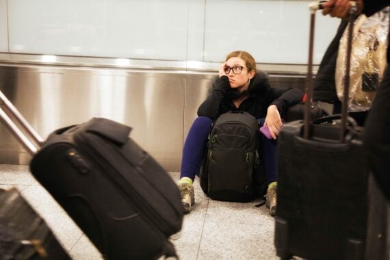 A woman sits on the airport floor, looking tired and resting her head on her hand, surrounded by luggage and travel bags as people walk by with suitcases.