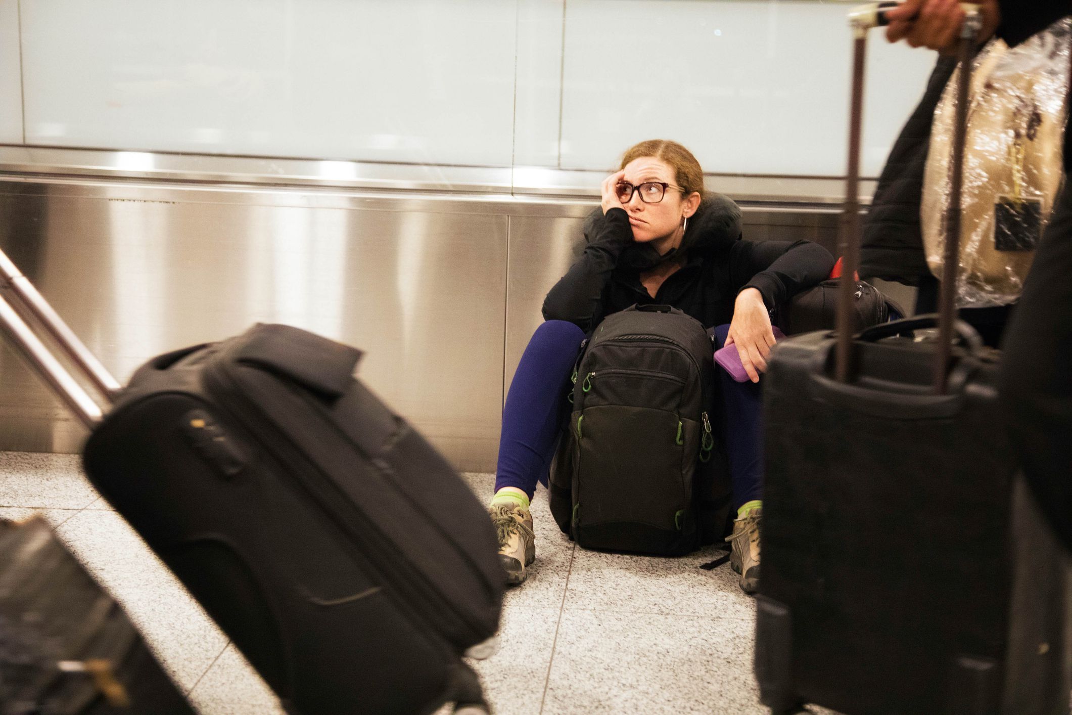 A woman sits on the airport floor, looking tired and resting her head on her hand, surrounded by luggage and travel bags as people walk by with suitcases.
