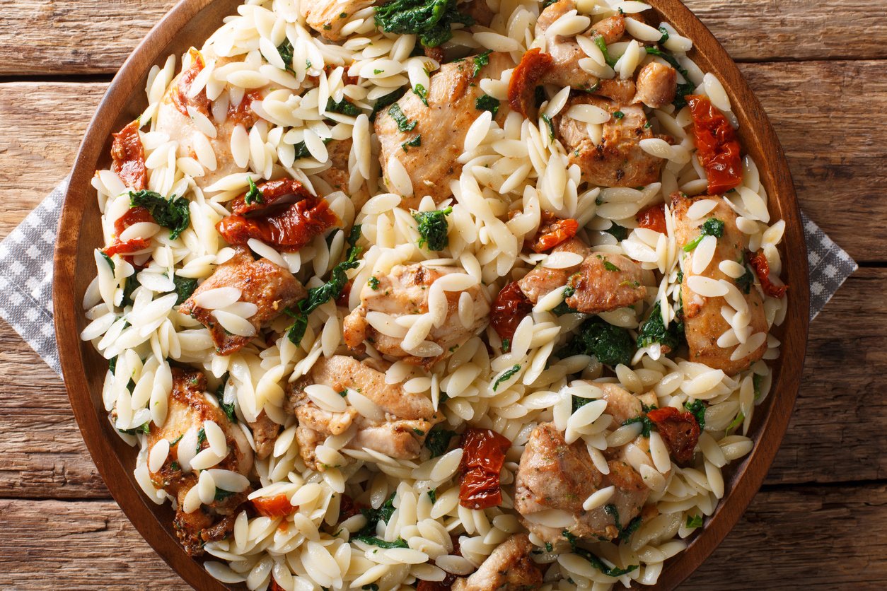 A wooden bowl filled with orzo pasta, pieces of cooked chicken, sun-dried tomatoes, and spinach, placed on a rustic wooden table with a gray checkered napkin underneath.