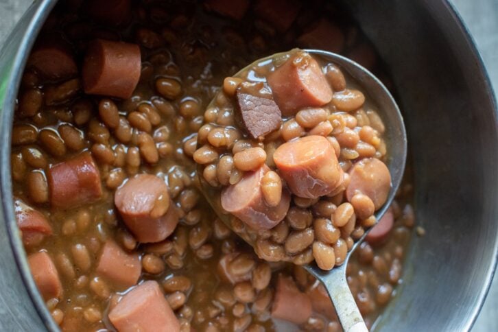 ladle of pork and beans with cut hot dog pieces over pot