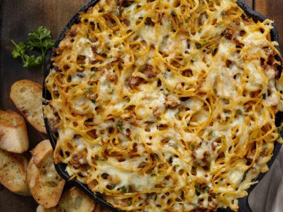 Baked spaghetti with melted cheese and meat in a cast-iron skillet, served with toasted bread slices.