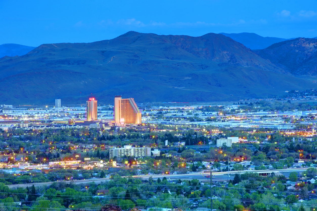 Cityscape at dusk with two illuminated tall buildings standing out among shorter structures, surrounded by greenery and roads, set against a backdrop of dark, tree-covered mountains.
