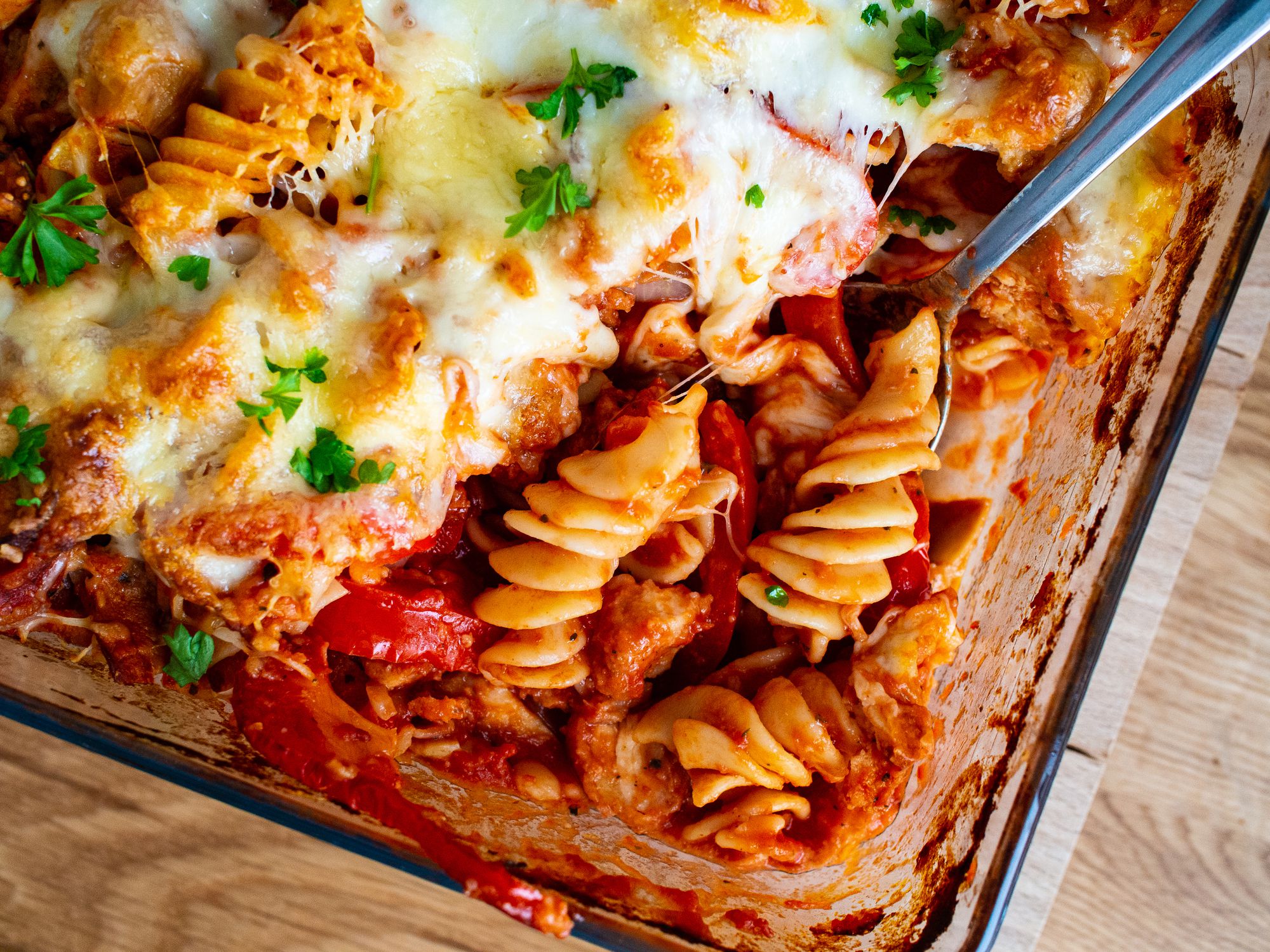 A close-up of a baked rotini pasta casserole with melted cheese, tomato sauce, and fresh parsley in a glass dish, with a serving spoon lifting out a portion.