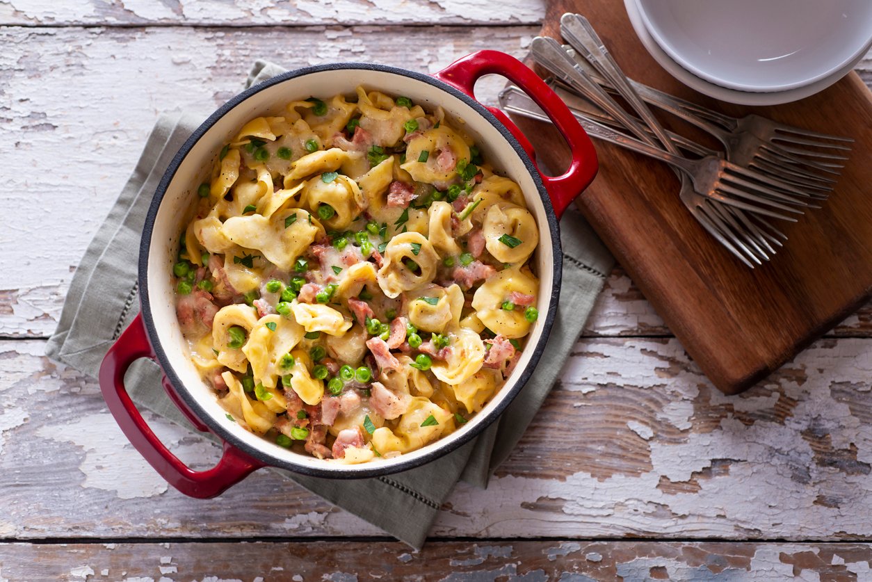 A red-handled pot filled with creamy tortellini pasta, diced ham, and green onions sits on a cloth napkin next to a wooden board with stacked plates and forks on a rustic white wooden table.