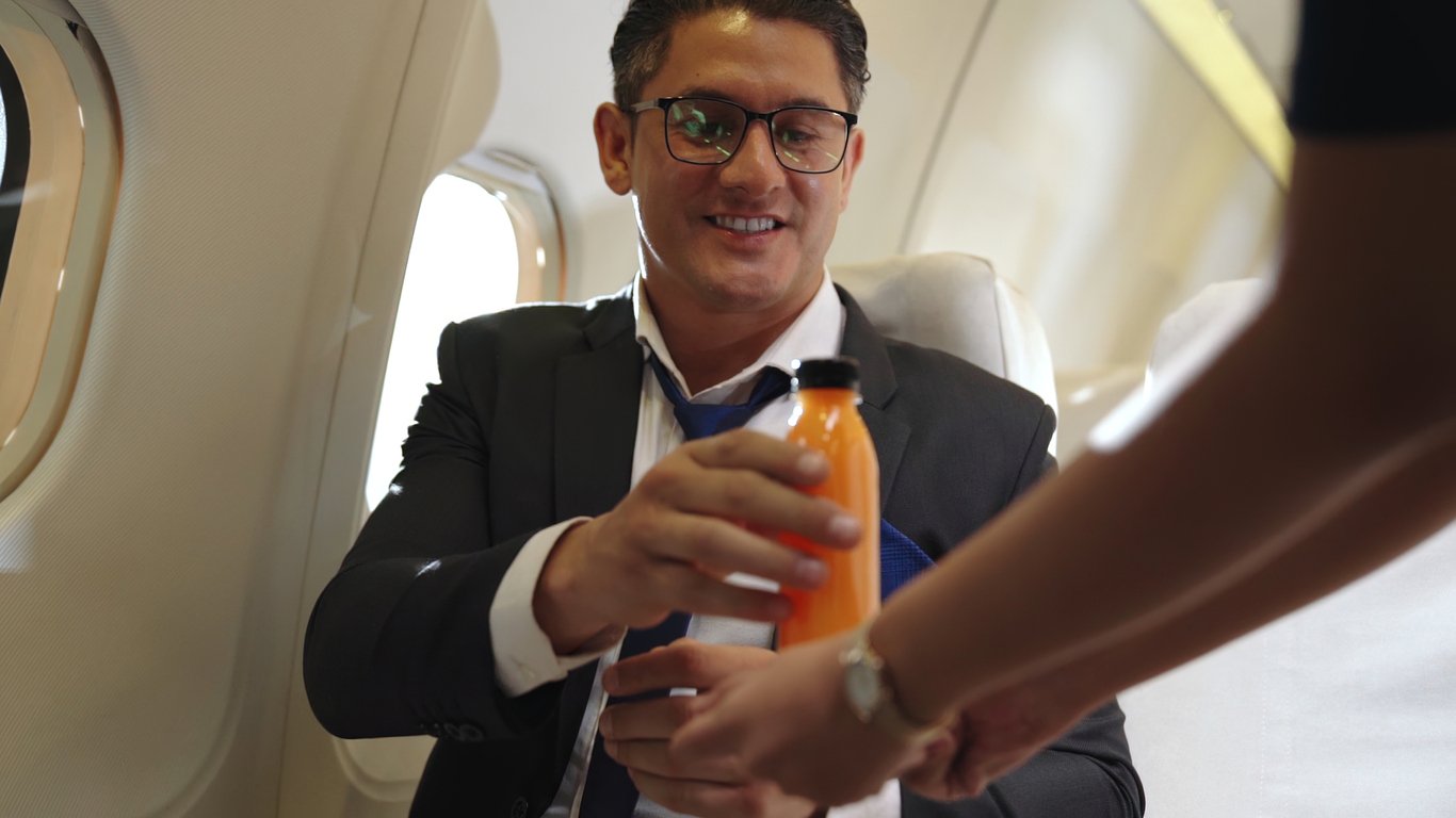 A man in a suit and glasses smiles as he receives a bottle of orange juice from a flight attendant on an airplane.