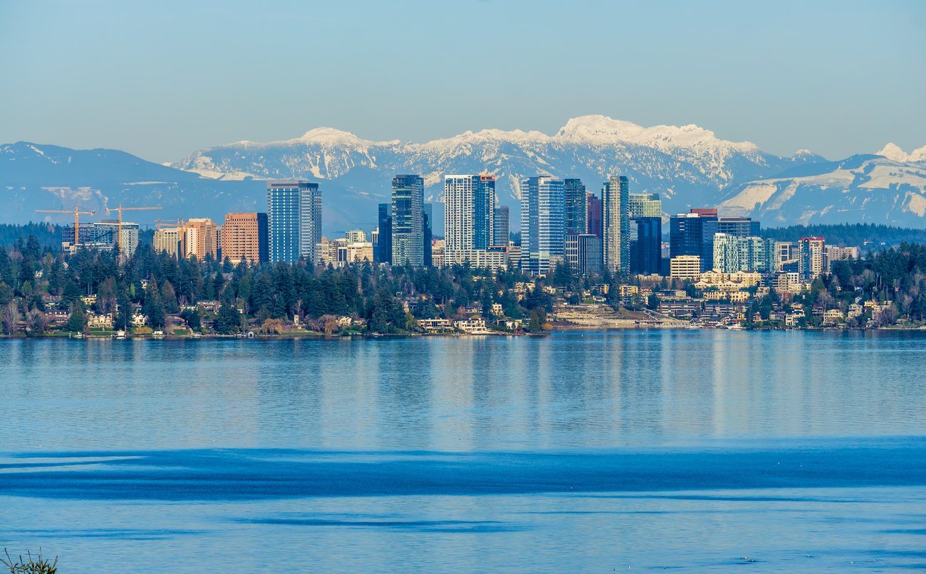 A city skyline with tall buildings is seen across a large body of water, with snow-capped mountains in the background under a clear blue sky. Dense green trees line the waterfront.