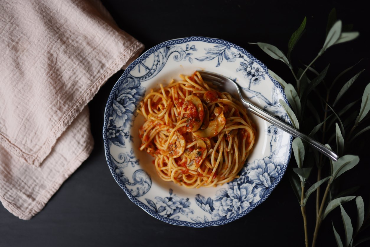 A decorative blue-and-white plate with spaghetti in tomato sauce and mushrooms, placed next to a fork, a light pink cloth, and green leaves on a dark surface.