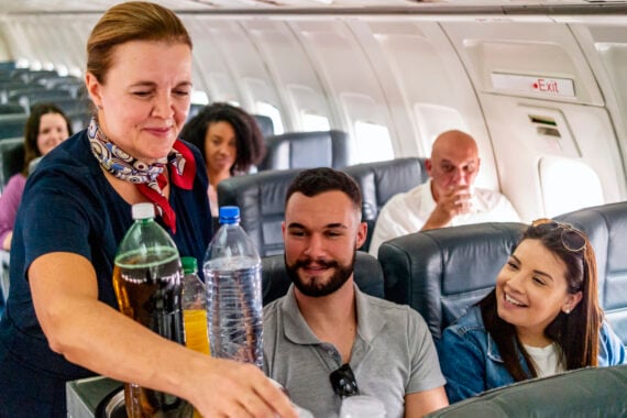 A flight attendant serves drinks to a smiling man and woman seated on an airplane, with other passengers visible in the background.