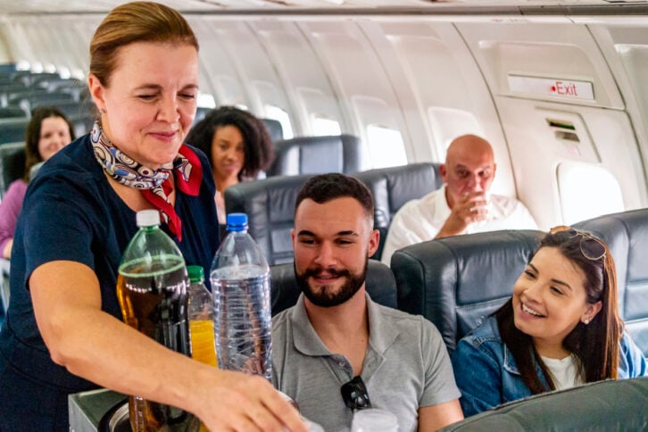 A flight attendant serves drinks to a smiling man and woman seated on an airplane, with other passengers visible in the background.