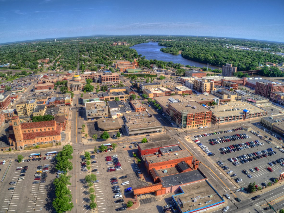 Aerial view of a mid-sized city with numerous buildings, parking lots, and tree-lined streets. A river flows in the background, surrounded by greenery under a clear blue sky.
