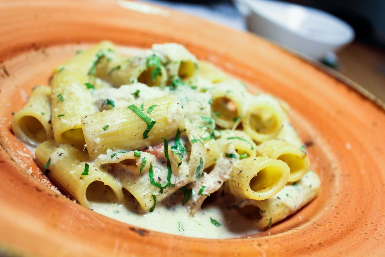 A plate of rigatoni pasta topped with creamy white sauce, grated cheese, chopped parsley, and pieces of meat, served on an orange ceramic dish.