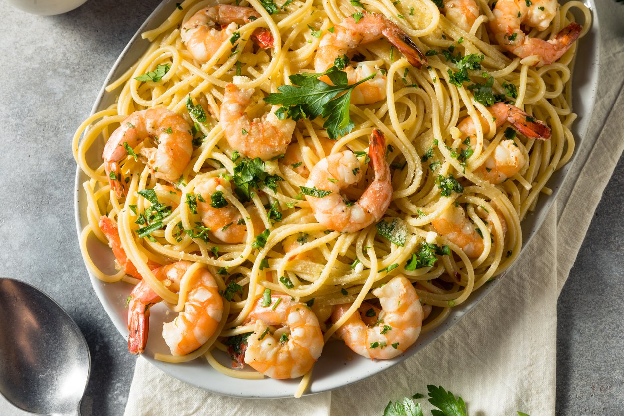 A plate of shrimp scampi pasta with linguine, garnished with chopped parsley. The dish features sautéed shrimp and appears lightly coated in a garlic butter sauce, served on a white plate with a napkin underneath.