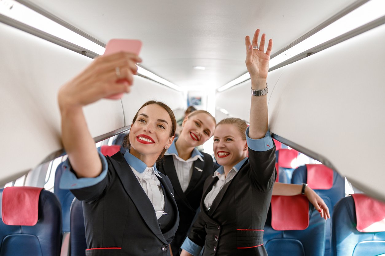 Three smiling flight attendants in uniform take a selfie inside an airplane cabin, standing in the aisle with passengers' seats and overhead compartments visible around them.