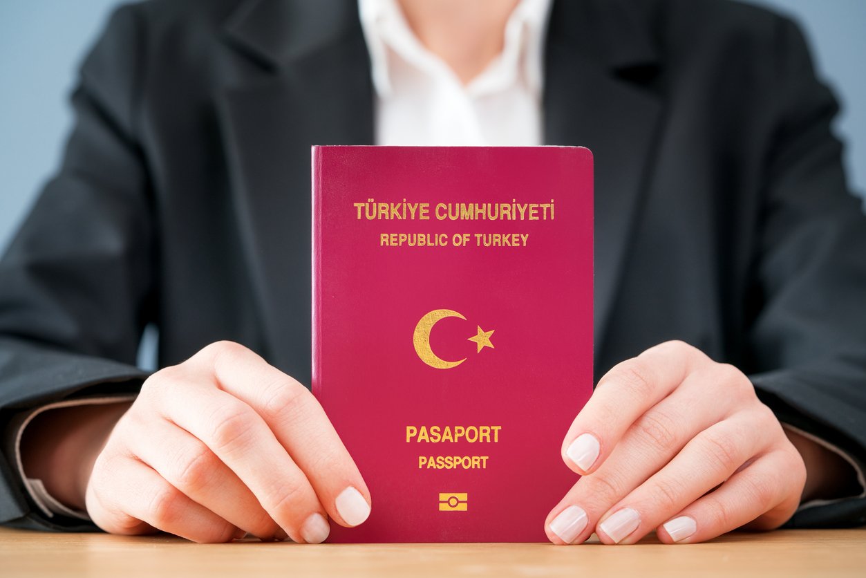 A person in a suit holds a maroon Turkish passport with both hands, showing the cover that displays the crescent moon, star, and gold lettering reading “Republic of Turkey Passport.”