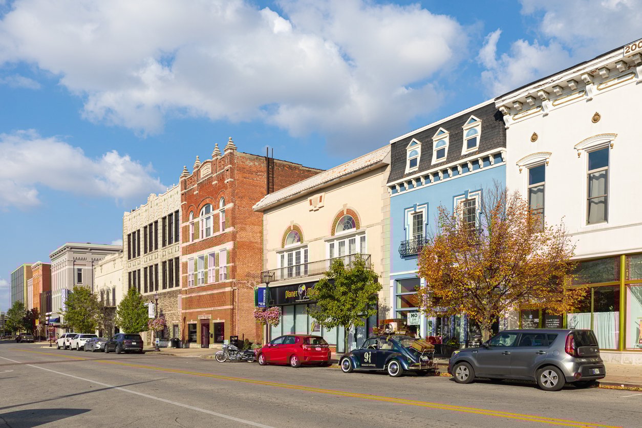 A sunny street scene shows historic downtown buildings with shops, parked cars, trees with autumn leaves, and a blue sky with scattered clouds.