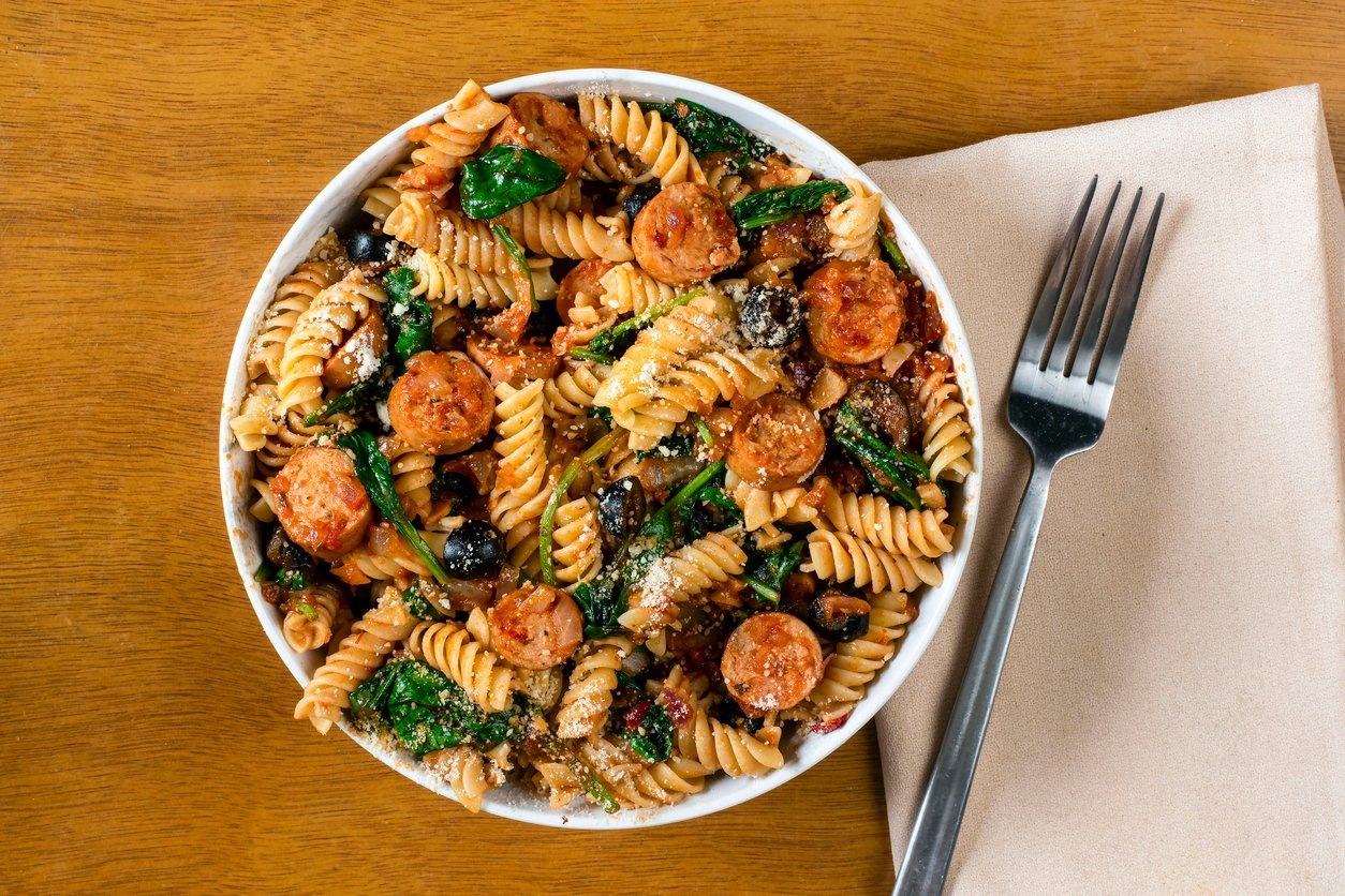 A bowl of rotini pasta with sliced sausage, black olives, spinach, and grated cheese sits on a wooden table next to a beige napkin and a fork.